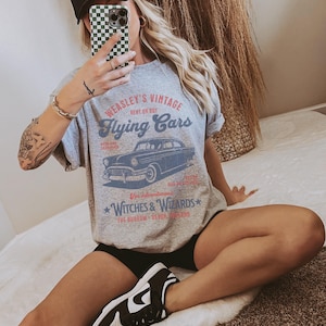 May include: Gray t-shirt with a vintage graphic of a flying car and the text "Weasley's Vintage Rent or Buy Flying Cars" and "Witches & Wizards The Burrow -  Devon, England".