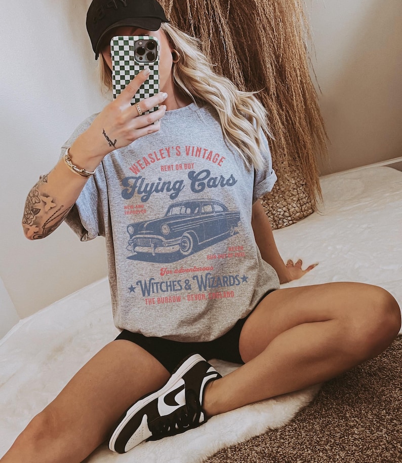 May include: Gray t-shirt with a vintage graphic of a flying car and the text "Weasley's Vintage Rent or Buy Flying Cars" and "Witches & Wizards The Burrow -  Devon, England".