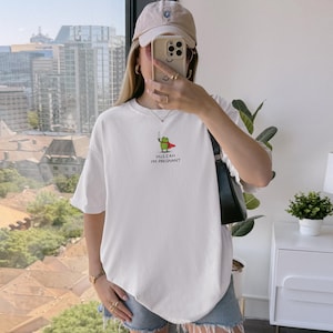 May include: White t-shirt with a graphic of a green frog wearing a red cape and holding a sword. The text "Huzzah I'm Pregnant" is below the frog. The shirt is short-sleeved and has a relaxed fit.