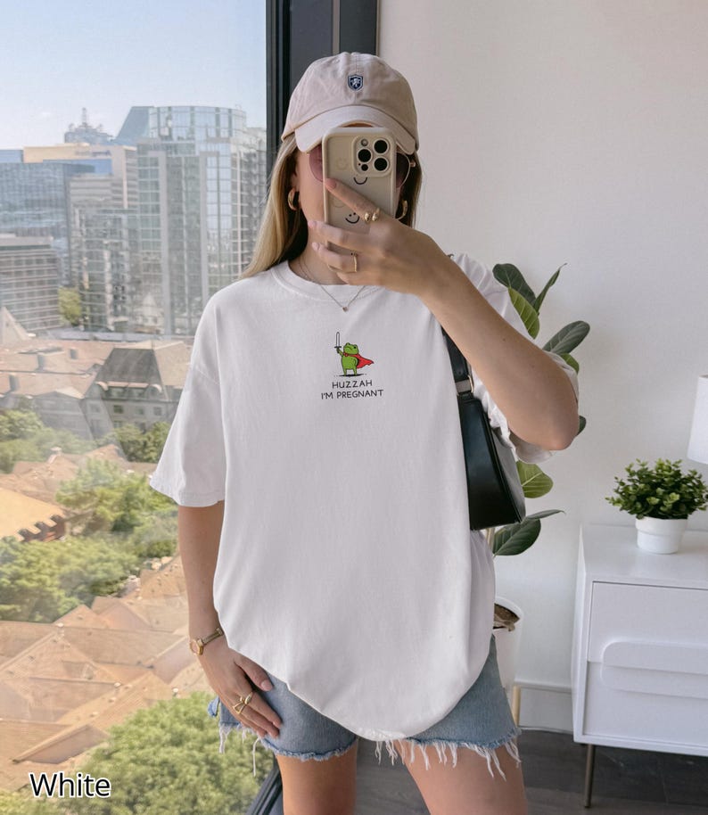 May include: White t-shirt with a graphic of a green frog wearing a red cape and holding a sword. The text "Huzzah I'm Pregnant" is below the frog. The shirt is short-sleeved and has a relaxed fit.