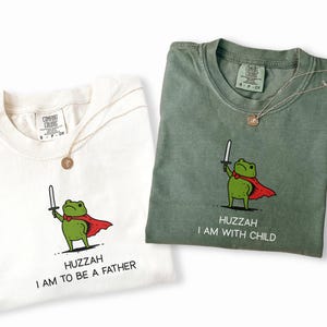 Puede incluir: Dos camisetas, marfil y verde musgo, cada una con un gr&aacute;fico de una rana verde con capa roja y espada. La camiseta marfil dice "HUZZAH I AM TO BE A FATHER", y la verde musgo dice "HUZZAH I AM WITH CHILD". Ambas tienen un collar dorado.
