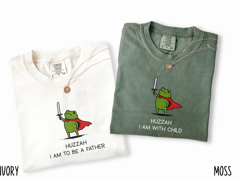 Puede incluir: Dos camisetas, marfil y verde musgo, cada una con un gr&aacute;fico de una rana verde con capa roja y espada. La camiseta marfil dice "HUZZAH I AM TO BE A FATHER", y la verde musgo dice "HUZZAH I AM WITH CHILD". Ambas tienen un collar dorado.