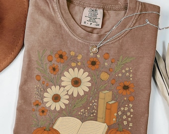 May include: A brown Comfort Colors t-shirt featuring a floral design with daisies, orange flowers, pumpkins, and books. A silver necklace rests on the shirt. The shirt is folded and ready to wear.
