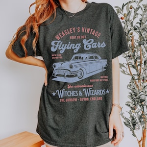 May include: A dark gray t-shirt with a vintage-style graphic of a flying car. The graphic says "Weasley's Vintage Flying Cars" and includes the text "Rent or Buy", "New and Improved", "For adventurous Witches & Wizards", "The Burrow - Devon, England", and "Never run out of fuel!"