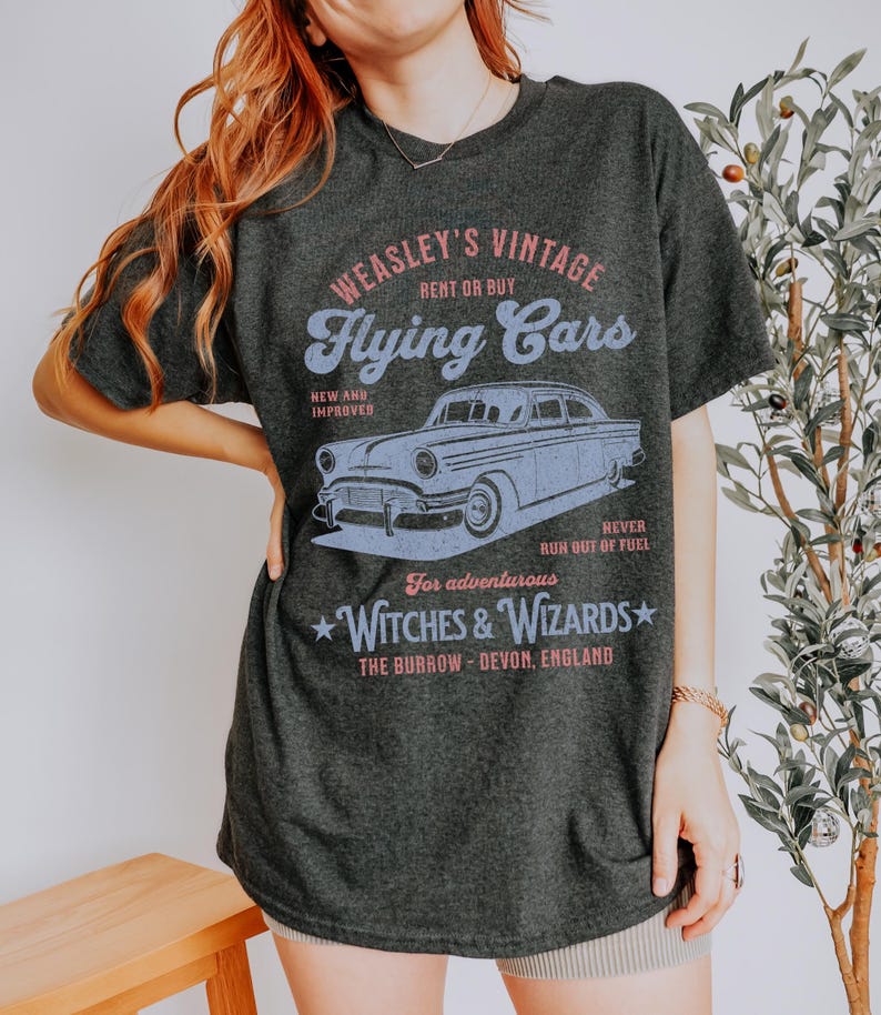 May include: A dark gray t-shirt with a vintage-style graphic of a flying car. The graphic says "Weasley's Vintage Flying Cars" and includes the text "Rent or Buy", "New and Improved", "For adventurous Witches & Wizards", "The Burrow - Devon, England", and "Never run out of fuel!"