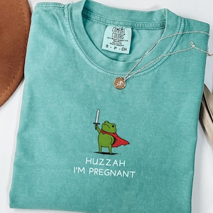 May include: Seafoam green Comfort Colors t-shirt with a graphic of a frog holding a sword and wearing a cape. The text "Huzzah I'm Pregnant" is below the frog. A silver necklace with a pendant is on the shirt.