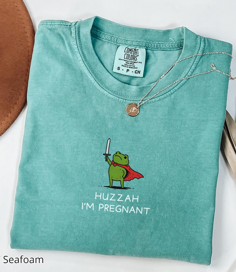 May include: Seafoam green Comfort Colors t-shirt with a graphic of a frog holding a sword and wearing a cape. The text "Huzzah I'm Pregnant" is below the frog. A silver necklace with a pendant is on the shirt.