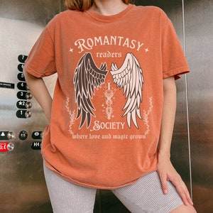 Puede incluir: Una camiseta naranja quemada con un gr&aacute;fico de alas y una espada. El texto "ROMANTASY readers SOCIETY where love and magic grows" est&aacute; impreso en la camiseta.
