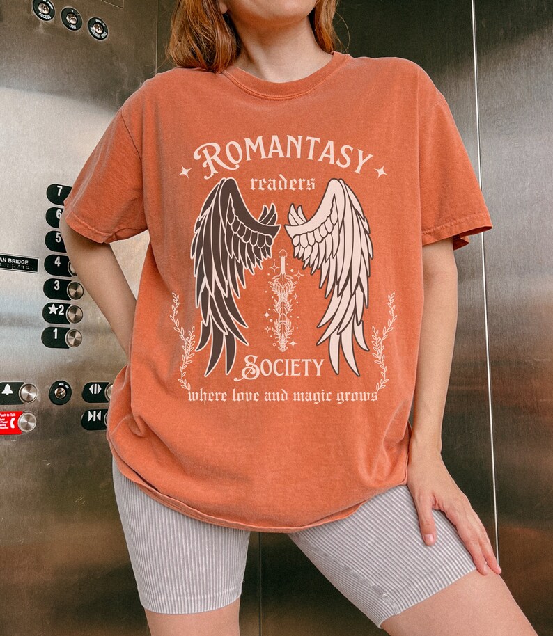 Puede incluir: Una camiseta naranja quemada con un gr&aacute;fico de alas y una espada. El texto "ROMANTASY readers SOCIETY where love and magic grows" est&aacute; impreso en la camiseta.