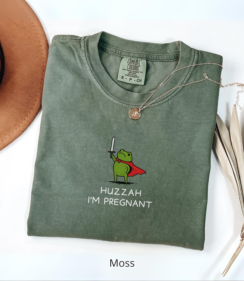 May include: Moss green t-shirt with a cartoon frog holding a sword and wearing a red cape. The text "Huzzah I'm Pregnant" is printed below the frog. A gold necklace and a brown hat are also visible.