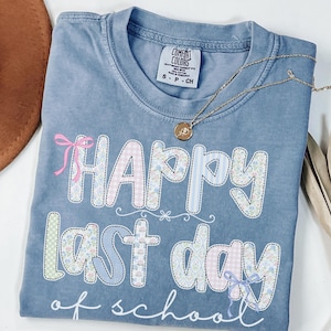 May include: Blue jean Comfort Colors t-shirt with the text "Happy Last Day of School" in a patchwork design. The shirt features pink, blue, and green patterns and a small bow. A gold necklace with a pendant is draped across the shirt.