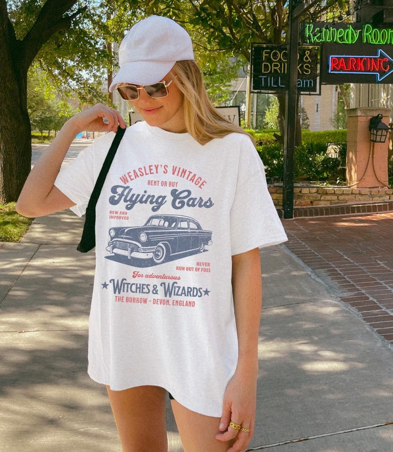 May include: White t-shirt with a vintage graphic design featuring a blue car and the text "WEASLEY'S VINTAGE RENT OR BUY Flying Cars NEW AND IMPROVED NEVER RUN OUT OF FUEL For adventurous WITCHES & WIZARDS THE BURROW - DEVON, ENGLAND"