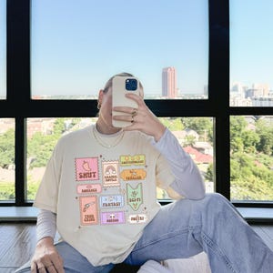 Może przedstawiać: Jasnobeżowy t-shirt z nadrukiem graficznym kolorowych znaczk&oacute;w przedstawiających r&oacute;żne gatunki literatury, w tym "Shut", "Romance", "Graphic Novels", "Thriller", "Fantasy", "Sci-Fi", "True Crime", "Horror" i "Poetry".