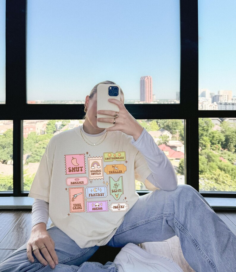 Może przedstawiać: Jasnobeżowy t-shirt z nadrukiem graficznym kolorowych znaczk&oacute;w przedstawiających r&oacute;żne gatunki literatury, w tym "Shut", "Romance", "Graphic Novels", "Thriller", "Fantasy", "Sci-Fi", "True Crime", "Horror" i "Poetry".