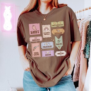 Może przedstawiać: Brązowy T-shirt z nadrukiem graficznym kolorowych znaczk&oacute;w przedstawiających r&oacute;żne gatunki literackie, w tym wsp&oacute;łczesny, shut, powieści graficzne, thriller, romans, fantasy, science fiction, true crime, horror i poezja.