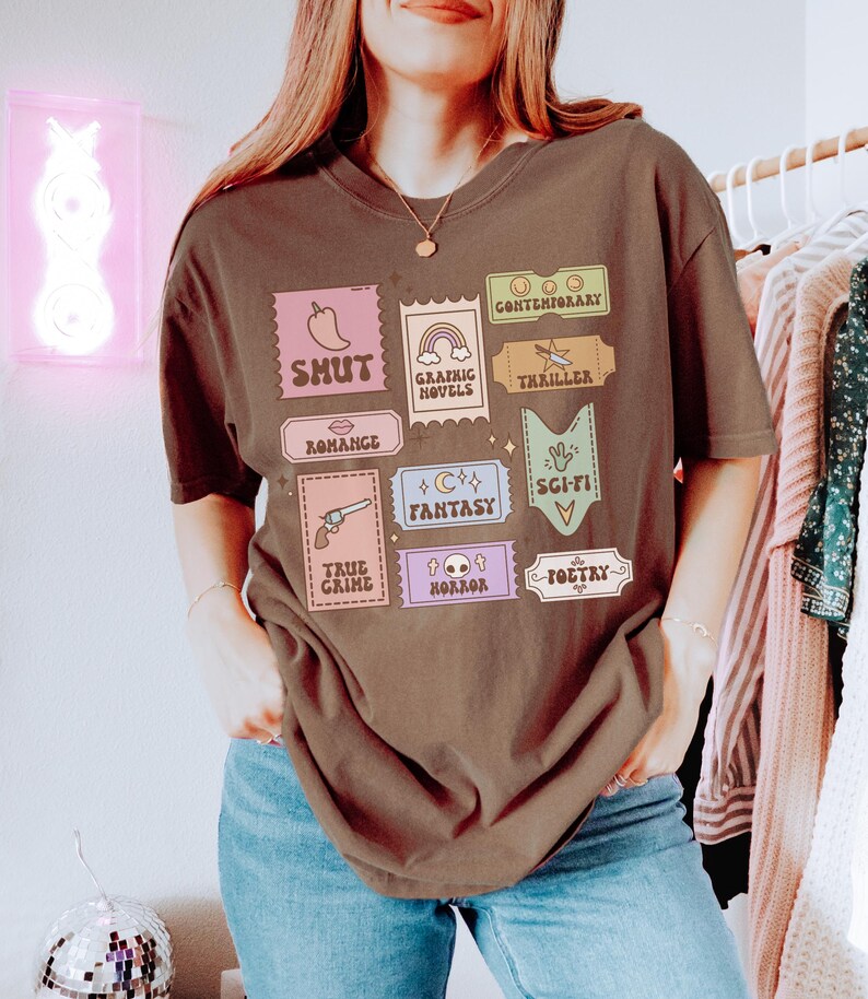 Może przedstawiać: Brązowy T-shirt z nadrukiem graficznym kolorowych znaczk&oacute;w przedstawiających r&oacute;żne gatunki literackie, w tym wsp&oacute;łczesny, shut, powieści graficzne, thriller, romans, fantasy, science fiction, true crime, horror i poezja.