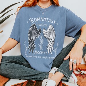 Puede incluir: Una camiseta azul con un gr&aacute;fico de alas de &aacute;ngel blancas y negras, una espada y el texto "ROMANTASY readers SOCIETY where love and magic grows".