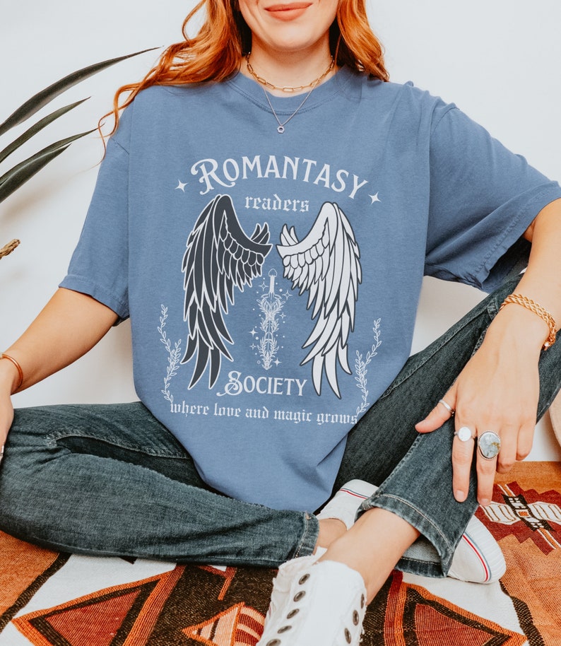 Puede incluir: Una camiseta azul con un gr&aacute;fico de alas de &aacute;ngel blancas y negras, una espada y el texto "ROMANTASY readers SOCIETY where love and magic grows".