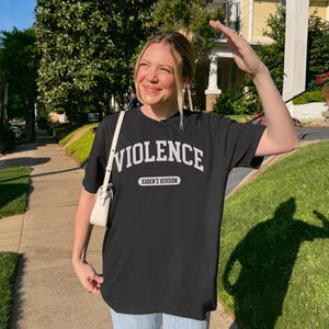Może przedstawiać: Czarny t-shirt z napisem "VIOLENCE" w białych literach i "XADEN'S VERSION" poniżej. Osoba nosi koszulkę na zewnątrz w słoneczny dzień.
