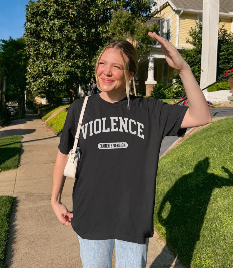 Może przedstawiać: Czarny t-shirt z napisem "VIOLENCE" w białych literach i "XADEN'S VERSION" poniżej. Osoba nosi koszulkę na zewnątrz w słoneczny dzień.