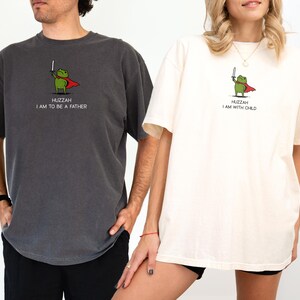 Puede incluir: Dos camisetas, una gris oscuro y otra crema, cada una con una rana de dibujos animados con capa y espada. La camiseta gris oscuro dice "Huzzah I am to be a father", y la camiseta crema dice "Huzzah I am with child".