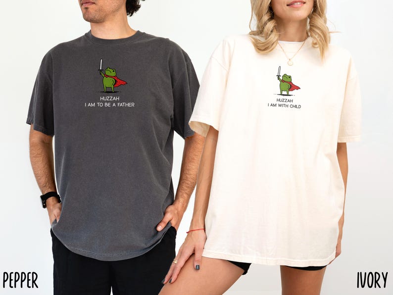 Puede incluir: Dos camisetas, una gris oscuro y otra crema, cada una con una rana de dibujos animados con capa y espada. La camiseta gris oscuro dice "Huzzah I am to be a father", y la camiseta crema dice "Huzzah I am with child".