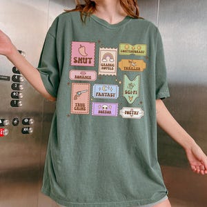 Może przedstawiać: Zielony, oversize'owy t-shirt z nadrukiem graficznym w postaci kolorowych, kwadratowych etykiet z r&oacute;żnymi gatunkami literatury. Gatunki te to: Shut, Romance, True Crime, Horror, Poetry, Fantasy, Sci-Fi, Thriller, Graphic Novels i Contemporary.