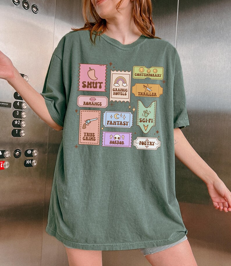 Może przedstawiać: Zielony, oversize'owy t-shirt z nadrukiem graficznym w postaci kolorowych, kwadratowych etykiet z r&oacute;żnymi gatunkami literatury. Gatunki te to: Shut, Romance, True Crime, Horror, Poetry, Fantasy, Sci-Fi, Thriller, Graphic Novels i Contemporary.