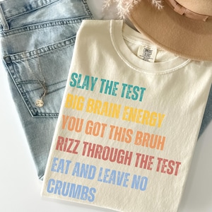 May include: Cream-coloured t-shirt with the text "SLAY THE TEST, BIG BRAIN ENERGY, YOU GOT THIS BRUH, RIZZ THROUGH THE TEST, EAT AND LEAVE NO CRUMBS" in various colours. The shirt is laid on top of light blue jeans, a necklace, and a tan hat.