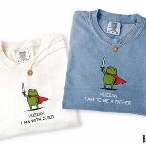 Puede incluir: Dos camisetas, una marfil y otra azul vaquero, cada una con una rana de dibujos animados sosteniendo una espada y usando una capa roja. La camiseta marfil dice "Huzzah I am with child" y la camiseta azul vaquero dice "Huzzah I am to be a father". Cada camiseta tiene un collar dorado.