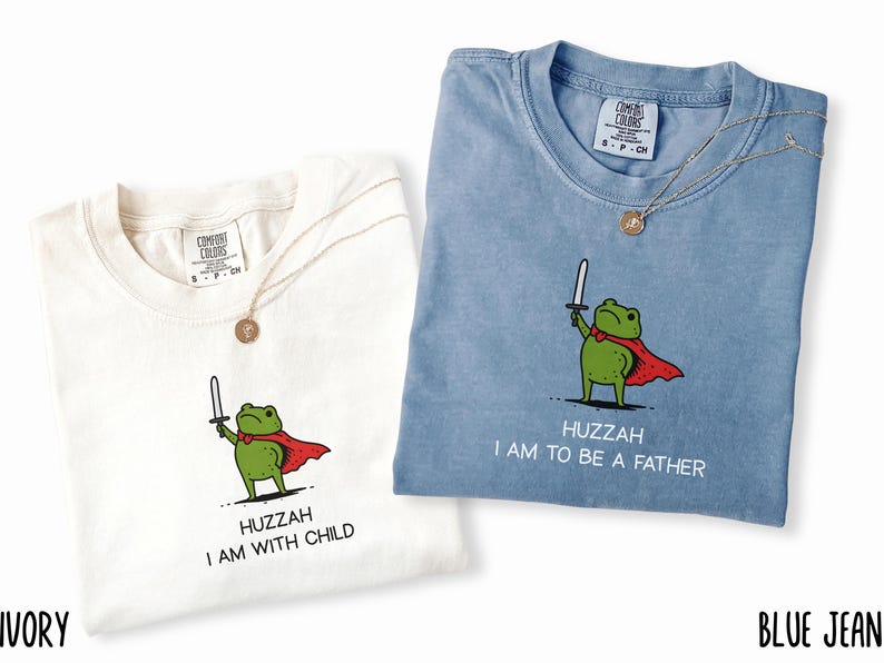 Puede incluir: Dos camisetas, una marfil y otra azul vaquero, cada una con una rana de dibujos animados sosteniendo una espada y usando una capa roja. La camiseta marfil dice "Huzzah I am with child" y la camiseta azul vaquero dice "Huzzah I am to be a father". Cada camiseta tiene un collar dorado.