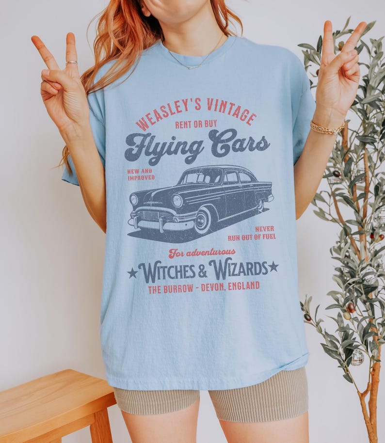 May include: Light blue t-shirt with a vintage graphic design featuring a black car and the text "WEASLEY'S VINTAGE Flying Cars RENT OR BUY NEW AND IMPROVED NEVER RUN OUT OF FUEL For adventurous WITCHES & WIZARDS THE BURROW - DEVON, ENGLAND"