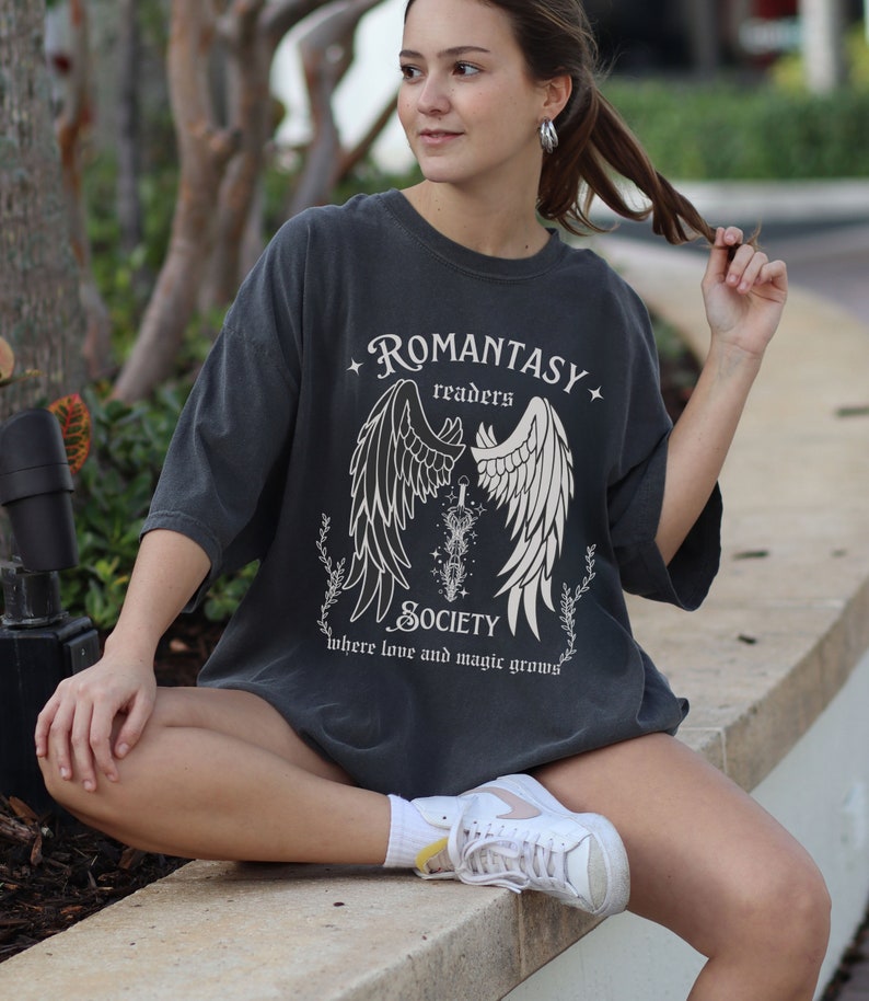 Puede incluir: Una camiseta gris oscuro de gran tama&ntilde;o con un dise&ntilde;o gr&aacute;fico de alas y el texto "ROMANTASY readers SOCIETY where love and magic grows"