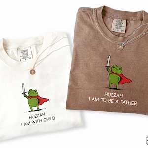Puede incluir: Dos camisetas Comfort Colors, marfil y espresso, cada una con un dise&ntilde;o de rana de dibujos animados. La camiseta marfil dice "HUZZAH I AM WITH CHILD", y la camiseta espresso dice "HUZZAH I AM TO BE A FATHER".