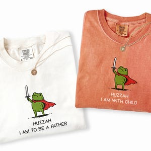 Puede incluir: Dos camisetas, una marfil y otra color &ntilde;ame, cada una con una rana de dibujos animados con capa y espada. La camiseta marfil dice "Huzzah I am to be a father". La camiseta color &ntilde;ame dice "Huzzah I am with child."