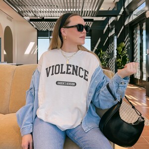 Może przedstawiać: Kremowa bluza z napisem "VIOLENCE" pogrubionymi, czarnymi literami. Poniżej słowa znajduje się napis "XADEN'S VERSION" mniejszą czcionką. Bluza jest noszona z dżinsową kurtką, niebieskimi dżinsami i czarną torebką.