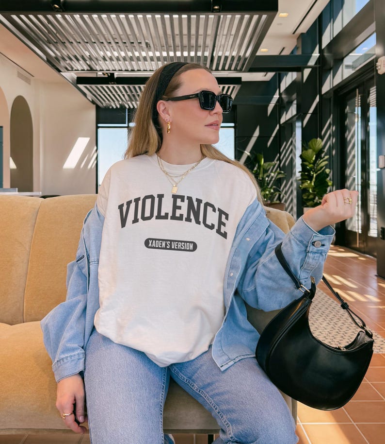 Może przedstawiać: Kremowa bluza z napisem "VIOLENCE" pogrubionymi, czarnymi literami. Poniżej słowa znajduje się napis "XADEN'S VERSION" mniejszą czcionką. Bluza jest noszona z dżinsową kurtką, niebieskimi dżinsami i czarną torebką.