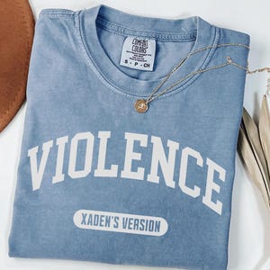 Może przedstawiać: Jasnoniebieska koszulka z napisem "VIOLENCE" dużymi białymi literami. Poniżej znajduje się tekst "XADEN'S VERSION" w białym prostokącie. Złoty naszyjnik z wisiorkiem spoczywa na koszulce.
