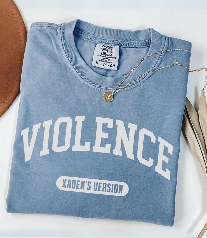 Może przedstawiać: Jasnoniebieska koszulka z napisem "VIOLENCE" dużymi białymi literami. Poniżej znajduje się tekst "XADEN'S VERSION" w białym prostokącie. Złoty naszyjnik z wisiorkiem spoczywa na koszulce.