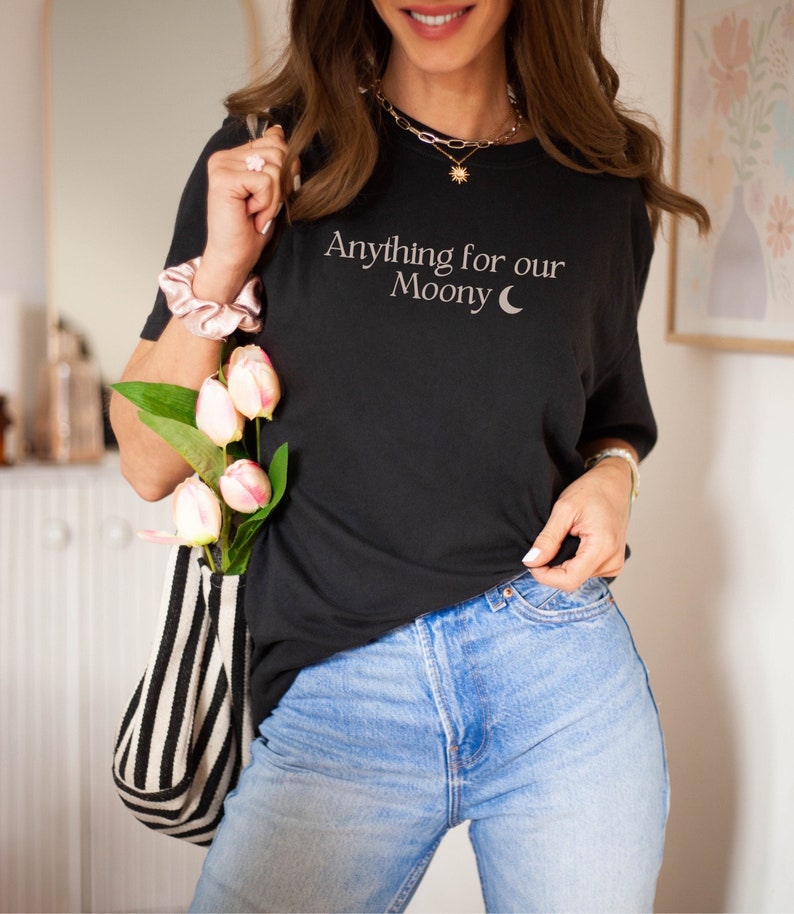 Może przedstawiać: Czarny T-shirt z napisem "Anything for our Moony" i grafiką rosnącego księżyca. Koszulkę nosi osoba z złotym naszyjnikiem i r&oacute;żową opaską na nadgarstku.