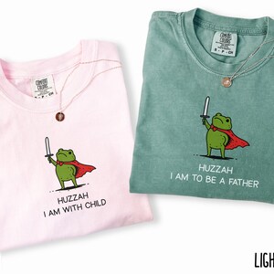 Puede incluir: Dos camisetas Comfort Colors, una rosa blossom y otra verde claro, con un dibujo de una rana. La rana sostiene una espada y lleva una capa roja. Los textos en las camisetas dicen "HUZZAH I AM WITH CHILD" y "HUZZAH I AM TO BE A FATHER".