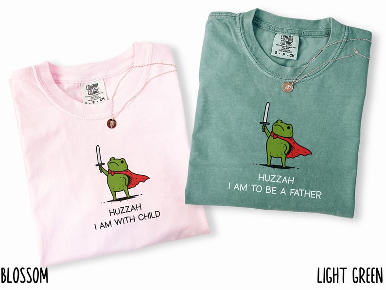 Puede incluir: Dos camisetas Comfort Colors, una rosa blossom y otra verde claro, con un dibujo de una rana. La rana sostiene una espada y lleva una capa roja. Los textos en las camisetas dicen "HUZZAH I AM WITH CHILD" y "HUZZAH I AM TO BE A FATHER".