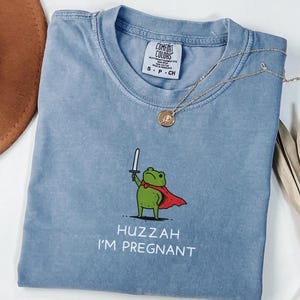 May include: Blue jean t-shirt with a graphic of a green frog wearing a red cape and holding a sword. The text "Huzzah I'm Pregnant" is below the frog. A gold necklace is draped across the shirt.