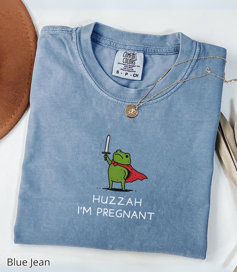 May include: Blue jean t-shirt with a graphic of a green frog wearing a red cape and holding a sword. The text "Huzzah I'm Pregnant" is below the frog. A gold necklace is draped across the shirt.