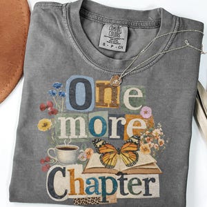 May include: Pepper-colored t-shirt with the words "One More Chapter" in a collage design. The graphic includes a butterfly, flowers, a coffee cup, and an open book. A silver necklace with a pendant rests on the shirt.