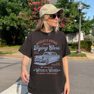 May include: Black t-shirt with a vintage graphic design featuring a flying car. The graphic says "Weasley's Vintage Flying Cars" and includes the text "Rent or Buy", "New and Improved", "Never Run Out of Fuel", "For adventurous Witches & Wizards", and "The Burrow - Devon, England".
