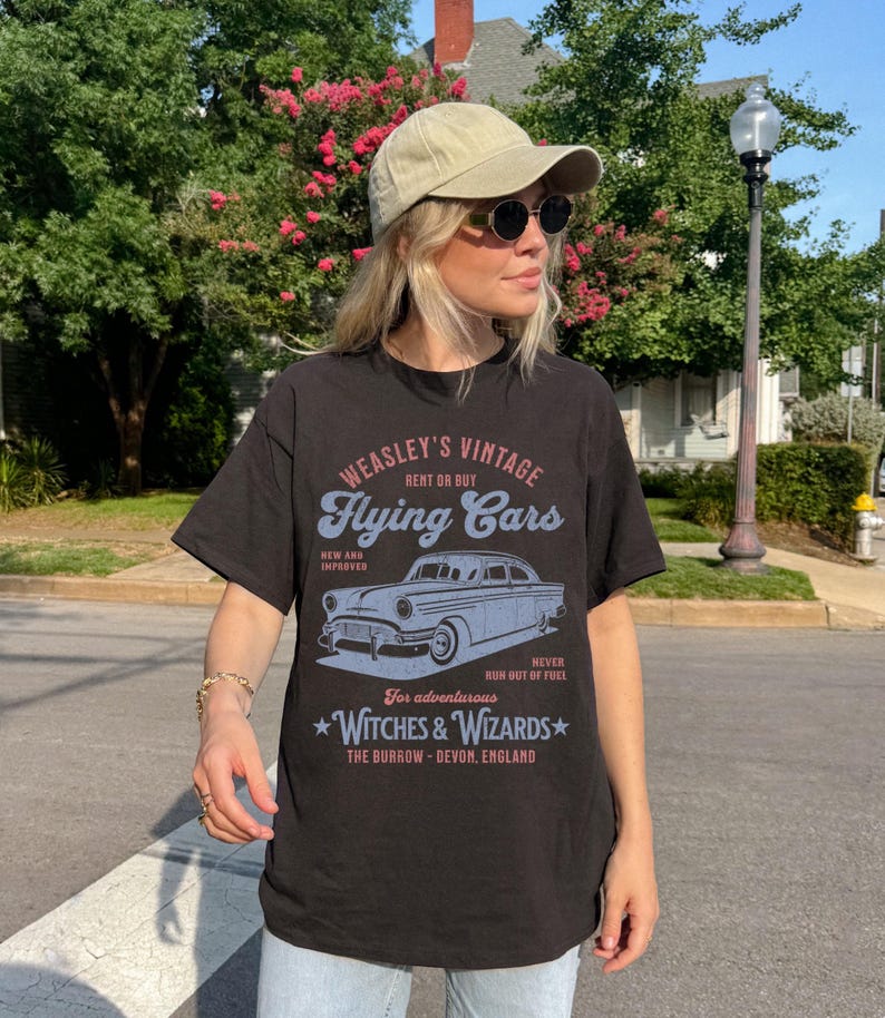 May include: Black t-shirt with a vintage graphic design featuring a flying car. The graphic says "Weasley's Vintage Flying Cars" and includes the text "Rent or Buy", "New and Improved", "Never Run Out of Fuel", "For adventurous Witches & Wizards", and "The Burrow - Devon, England".