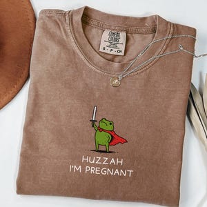 May include: Espresso-colored t-shirt with a crew neck. The shirt features a cartoon frog wearing a red cape and holding a sword, with the text "Huzzah I'm Pregnant". A silver necklace with a pendant rests on the shirt.