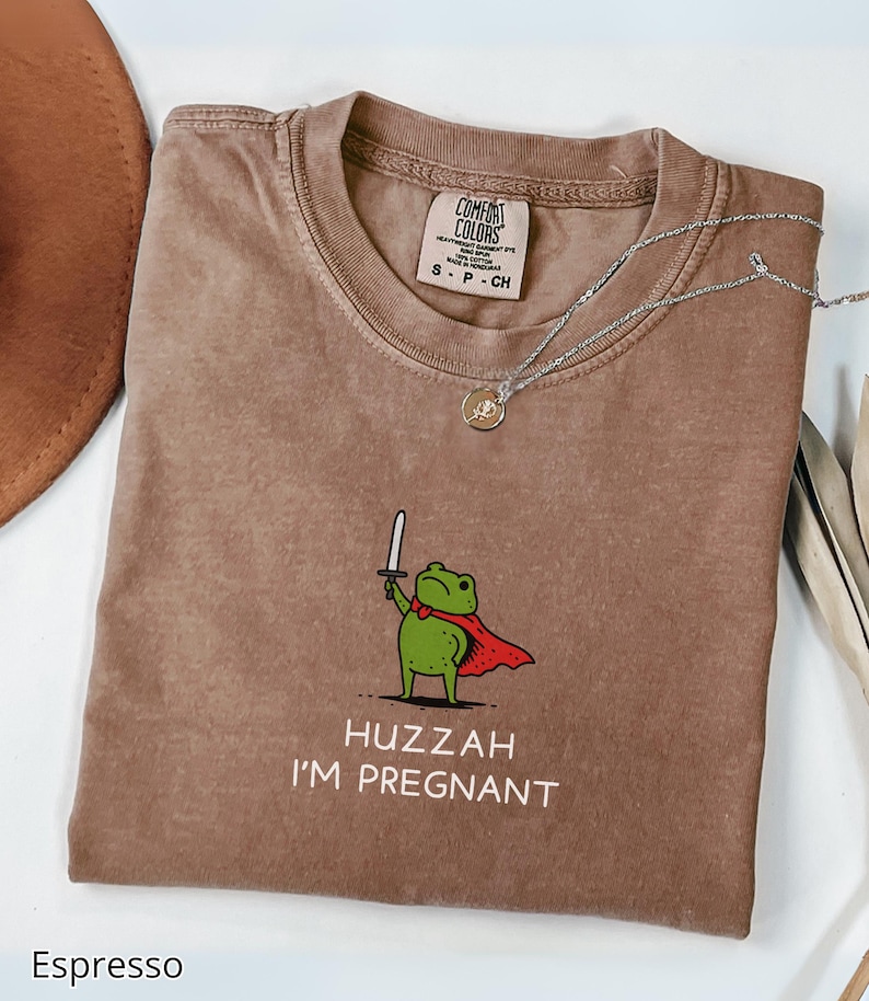 May include: Espresso-colored t-shirt with a crew neck. The shirt features a cartoon frog wearing a red cape and holding a sword, with the text "Huzzah I'm Pregnant". A silver necklace with a pendant rests on the shirt.