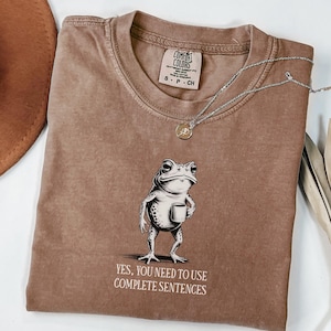 May include: Espresso-coloured t-shirt featuring a frog graphic with a cup and the text "YES, YOU NEED TO USE COMPLETE SENTENCES." A silver necklace with a small pendant lies on the shirt. The shirt has a crew neck and short sleeves.